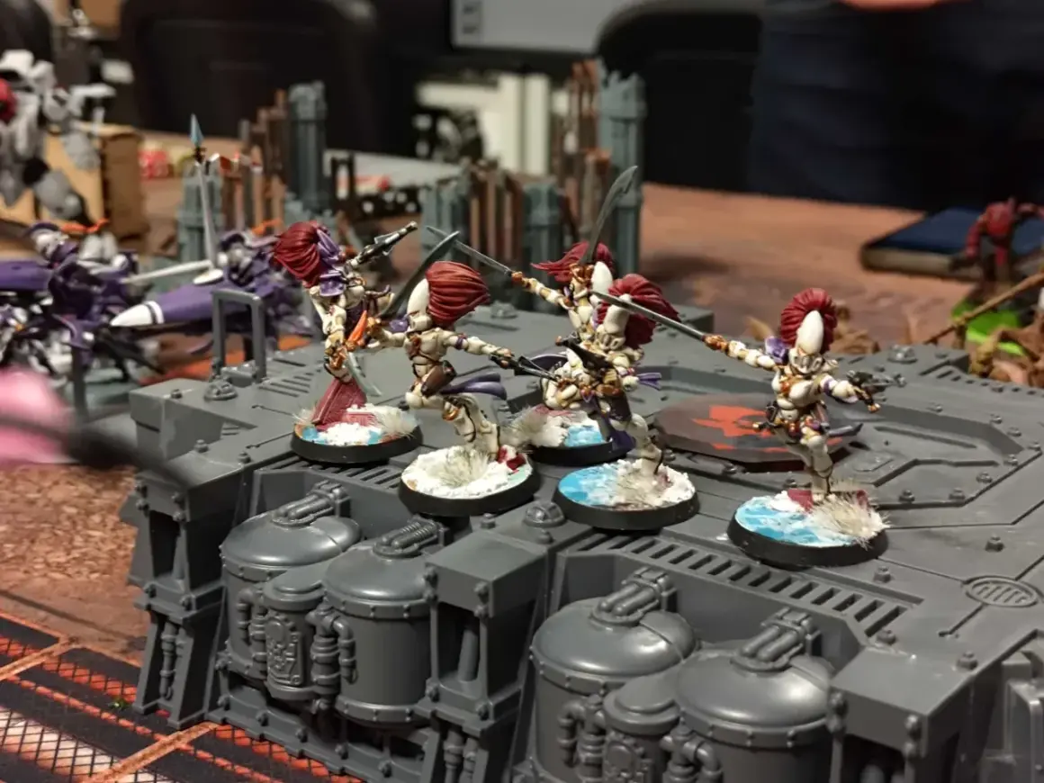Figurki wojowniczek Eldarów, jednej z frakcji Warhammer 40k, szykują się do walki na szarej platformie.